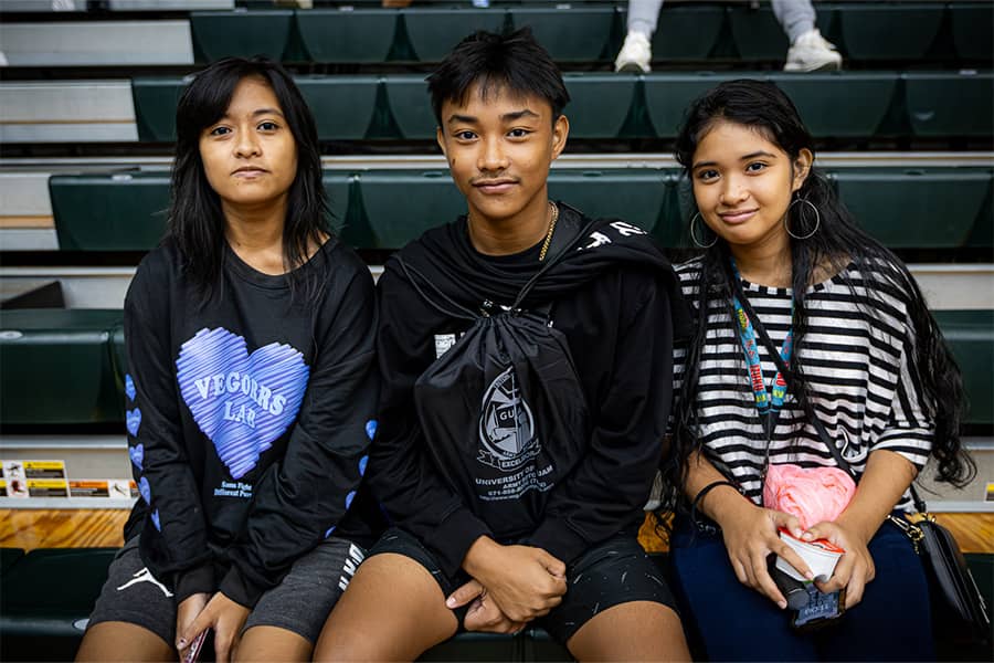Three students, siblings, pose for a photo sitting at the UOG Fieldhouse bleachers