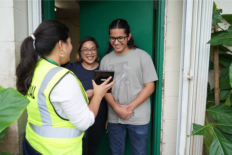 A woman in a neon vest holds a tablet up explaining something to a couple of UOG students