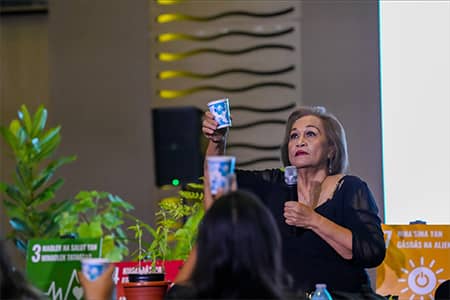 Mama Lou, with a microphone in hand, holds cups in the air with audience members of G3 workshop.