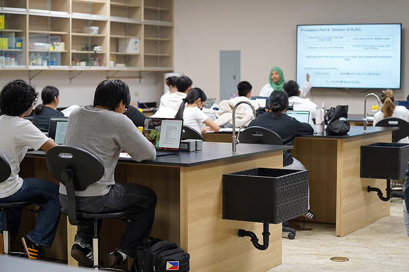 A chemistry professor in a lab coat at the front of a lab by a screen speaking to a classroom of students seated at new chemistry lab benches.