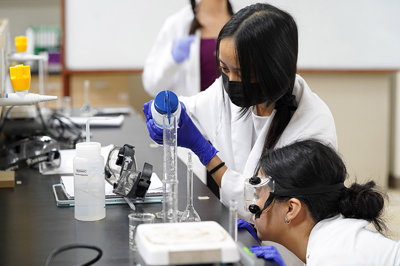 Two students in lab coats add a liquid to a beaker at a chemistry lab bench station.