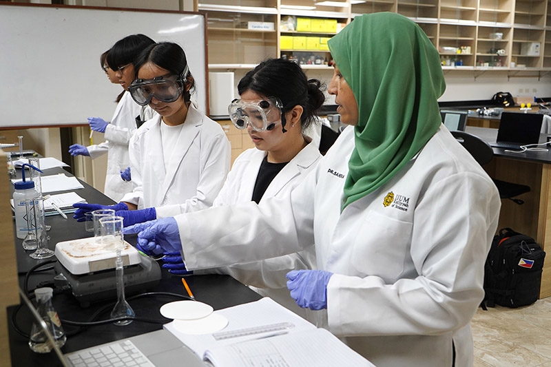 A chemistry professor points to a beaker while two students in goggles and lab coats look on.